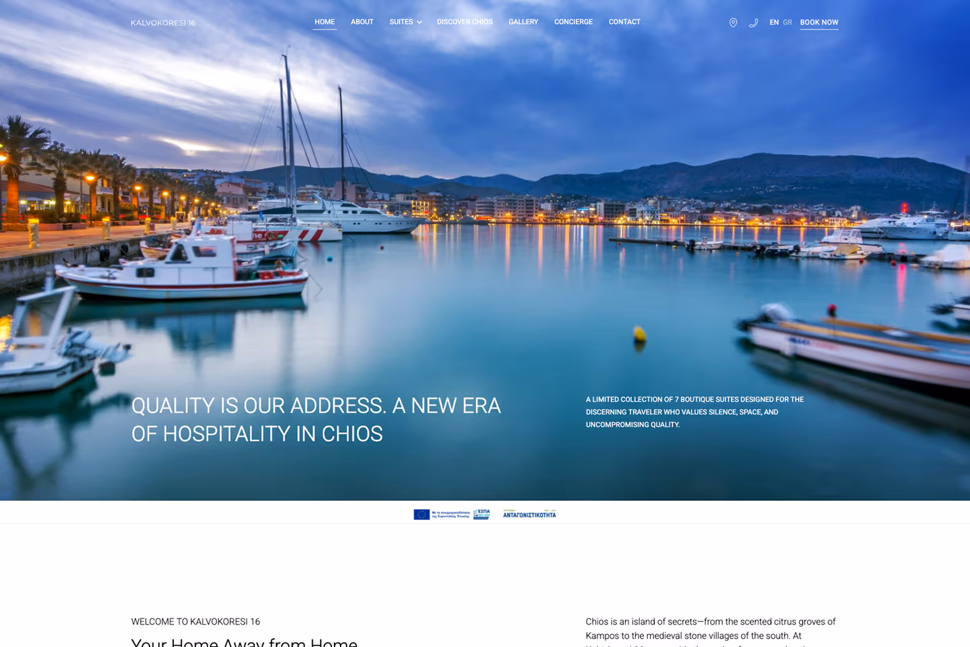Boats docked in calm harbor waters with a town and mountains under a cloudy evening sky.