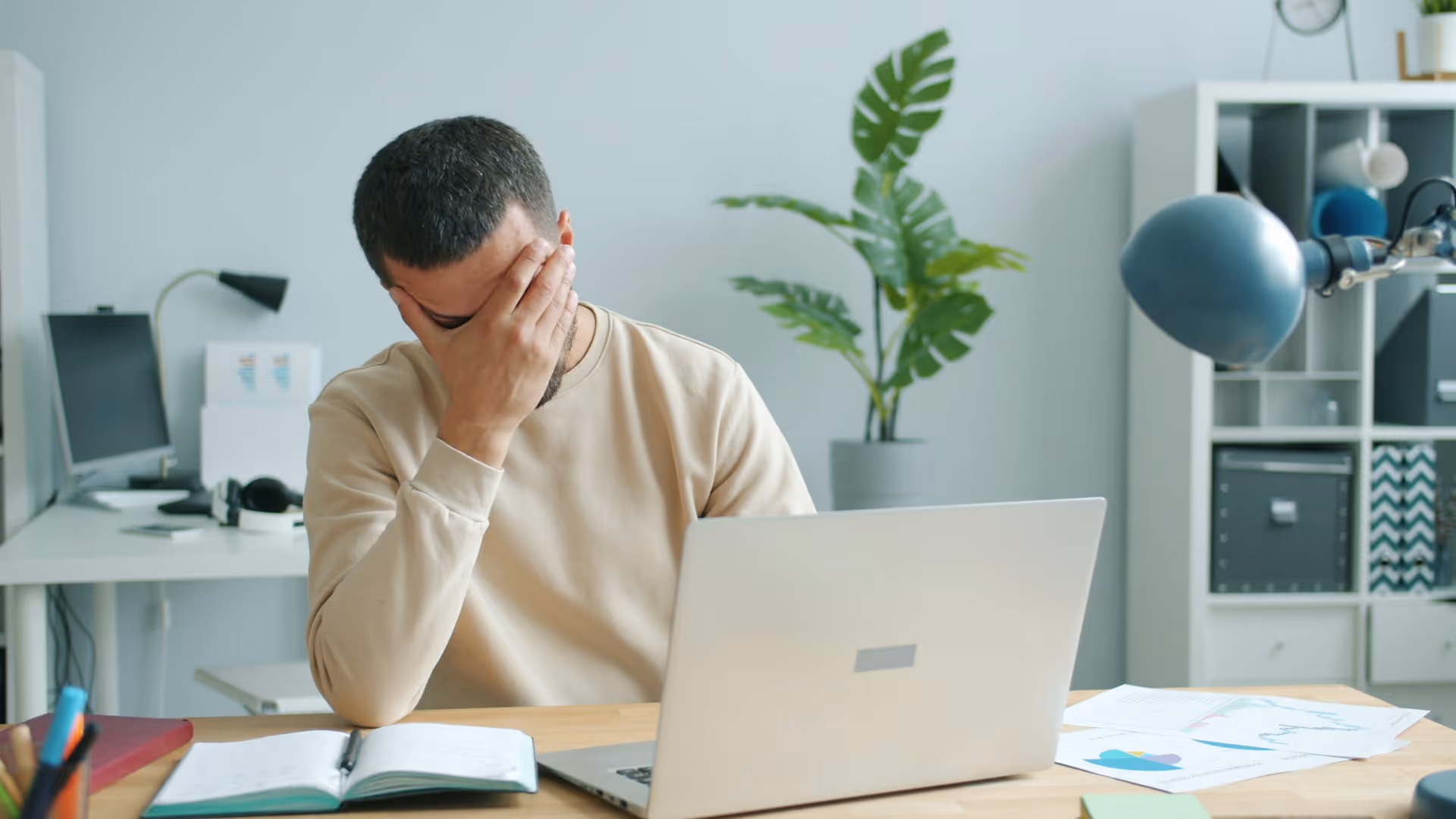 Frustrated man sitting at desk with laptop, notebook, and papers in a modern office.
