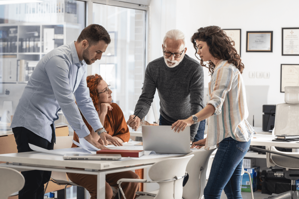 Four coworkers gathered around a table, discussing documents and a laptop in a bright office.