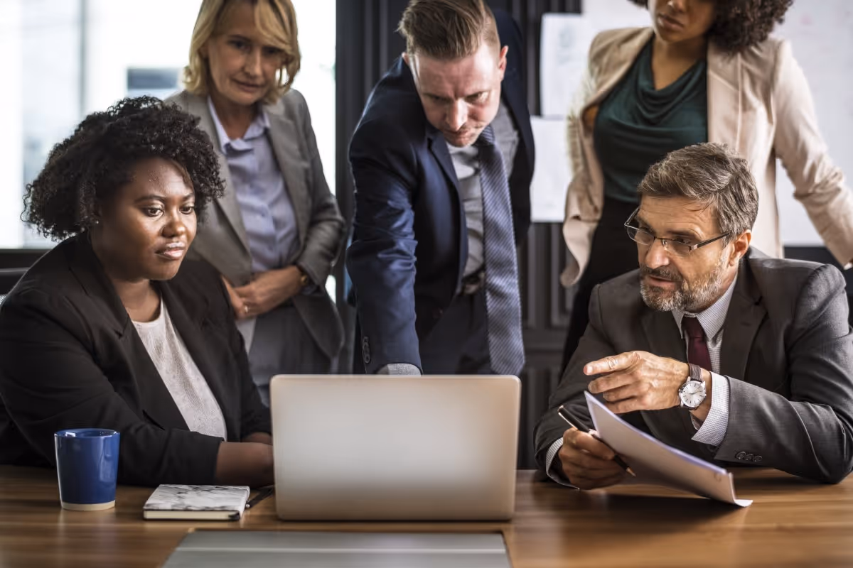 Five diverse businesspeople gathered around a laptop in a meeting room discussing documents.